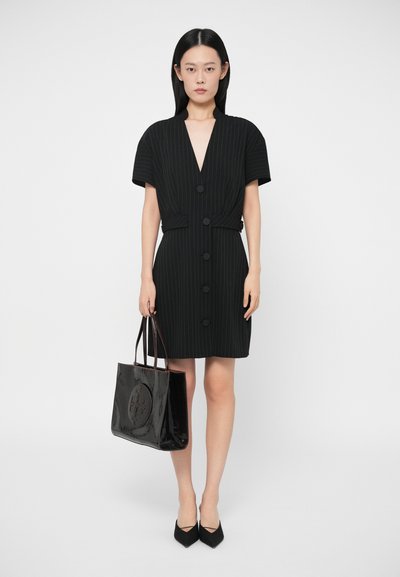 Kobieta w czarnej sukience w paski z dużymi guzikami, trzymająca czarną, błyszczącą torebkę oraz nosząca czarne, szykowne szpilki, stojąca na białym tle.