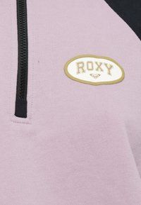 Svetlofialový pulóver s čiernym zipsom a bielou oválnou nášivkou s nápisom "ROXY" v zlatom písmene. Mäkká textúra, športový dizajn.