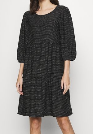 Mujer con vestido negro hasta la rodilla con rayas horizontales blancas y mangas abullonadas tres cuartos, sobre un fondo claro y liso.