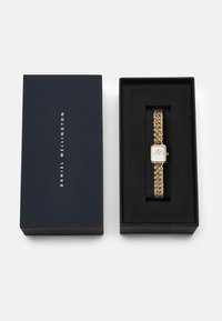 Montre en or avec un cadran blanc rectangulaire et un bracelet en chaîne épais, présentée dans une boîte bleu foncé avec du texte doré. Intérieur en velours noir.