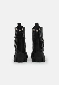 Botas de plataforma negras, gruesas y con cremallera trasera, tiradores de marca y correas con hebillas a los lados, sobre un fondo blanco liso.