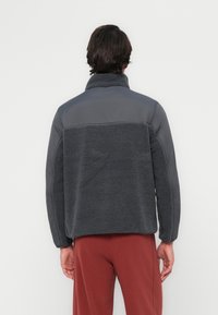 Donkergrijze fleecejas met een glad bovenpaneel en een gestructureerd lijf. Voorzien van een hoge kraag en lange mouwen. Eenvoudig ontwerp zonder zichtbare branding.