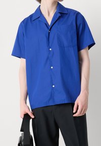 Koszula z krótkim rękawem i kołnierzykiem, wykonana z jednolitej, jasnoniebieskiej bawełny. Posiada jedno kieszonkę i białe guziki, o luźnym kroju. Noszona z czarnymi spodniami.
