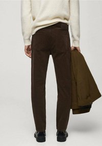 Bruine corduroybroek met een slim fit, voorzien van twee achterzakken, gedragen met een crèmekleurige trui en een groene jas vastgehouden. Zwarte schoenen zichtbaar.