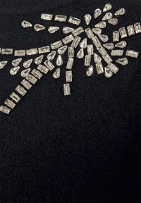 Pedrería de cristal transparente en forma rectangular y de lágrima, dispuesta en un patrón en forma de explosión sobre un fondo de tela negra.