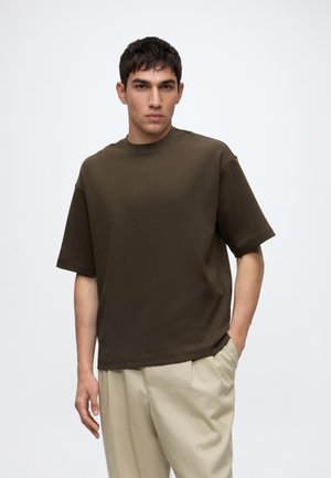 Jeune homme portant une chemise oversize à manches courtes marron foncé et un pantalon beige, une main dans la poche, sur un fond uni.