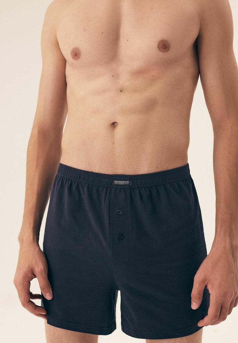 Ciemnoniebieskie bawełniane bokserki z elastycznym pasem, z dwoma guzikami z przodu i gładką teksturą. Model nie ma na sobie koszuli.