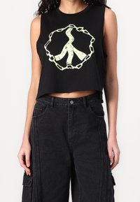 Sort cropped tank top med et lysende grønt fredssymbol omgivet af et kædedesign. Båret med højtaljede sorte denimbukser.