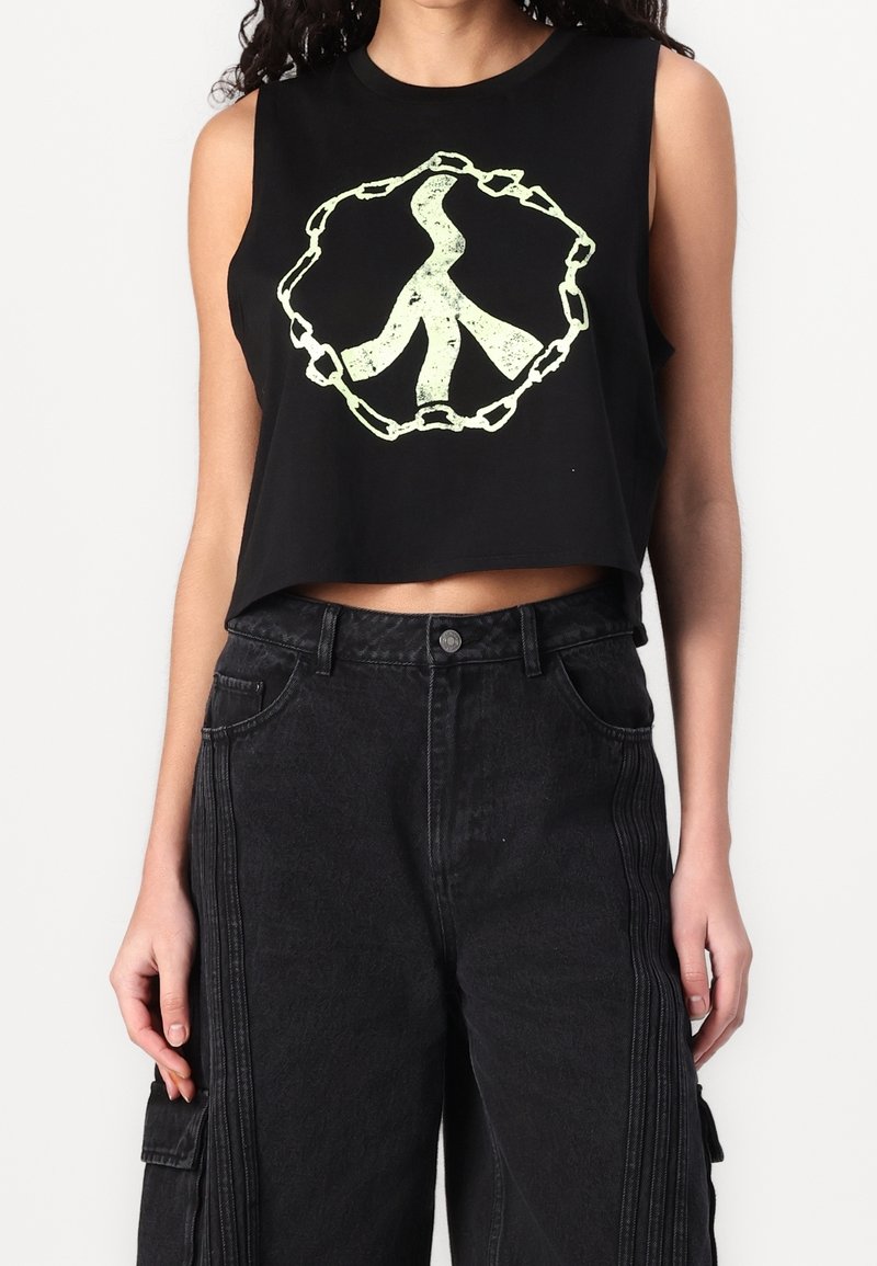 Sort cropped tank top med et lysende grønt fredssymbol omgivet af et kædedesign. Båret med højtaljede sorte denimbukser.