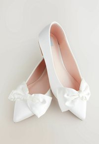 Ballerines satinées blanches avec un bout pointu et un grand nœud sur chaque chaussure. L'intérieur est rose avec une texture lisse.
