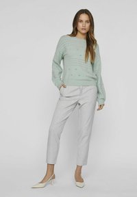 Světle zelený pletený svetr s perforovanými vzory, kombinovaný se světle šedými úzkými kalhotami a krémovými lodičkami na podpatku.