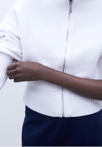 Vit dragkedjetröja med ribbad struktur vid fållen och manschetterna, bärs över marinblå byxor. Tyget ser mjukt och lätt ut.