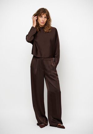 Vrouw met golvend haar, die een donkerbruine top met lange mouwen en wijde broek met bruine puntige schoenen draagt, staand tegen een witte achtergrond.