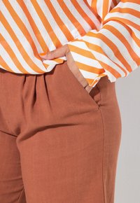 Pruhovaná blůza v bílé a oranžové barvě, s dlouhými rukávy, nošená přes zářivě hnědé skládací kalhoty. Textura se zdá být měkká a lehká.