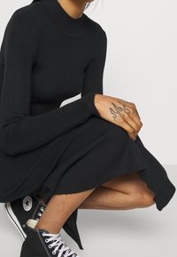 Personne portant une robe noire côtelée à manches longues et des baskets montantes Converse noires, montrant un tatouage de main avec un motif de flamme.