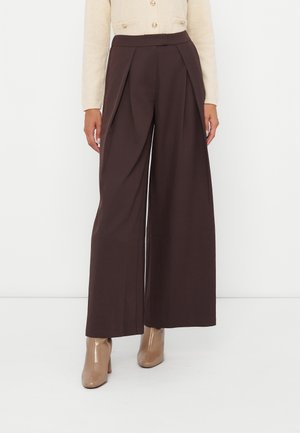 Femme portant un pantalon large marron foncé, un pull boutonné beige et des bottines beige, debout devant un fond blanc.
