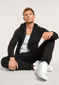 Haut à capuche noir zippé avec des accents rouges, associé à un jogging noir assorti et des baskets blanches. Le tissu semble doux et confortable.