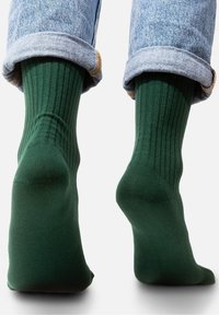 Zielone prążkowane skarpetki o dopasowanym kroju, wykonane z gładkiego materiału. Zawinięte z jasnymi dżinsami, eksponując teksturę i kontrast kolorów.