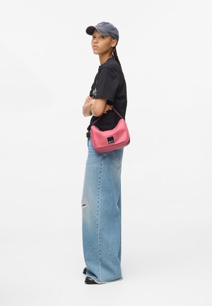 Jonge vrouw in een zwarte pet en T-shirt met brede, gescheurde jeans houdt een kleine roze handtas over haar arm.