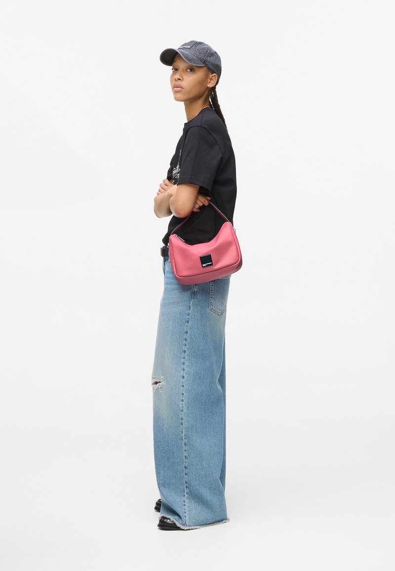 Ung kvinde iført en sort kasket og T-shirt med vide, ripped jeans bærer en lille pink håndtaske over armen.