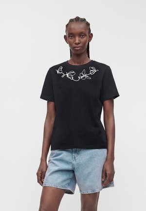 Kvinde iført en sort t-shirt med hvide sommerfugle-linjedesigns på brystet, kombineret med lyseblå denimshorts, stående mod en ensfarvet baggrund.