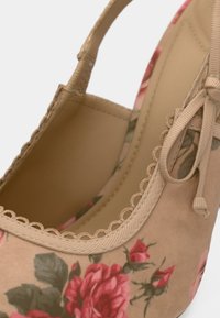 Zapato de tacón alto beige con un patrón floral en tonos rosas, que presenta un borde festoneado y un detalle de cinta texturizada en el lateral.
