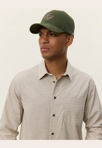 Grønn caps med buet skjerm, har brodert logo. Bært med en beigefarget rutete skjorte med knapper, som viser en subtil tekstur.