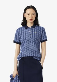 Jeune femme portant un polo bleu à motifs et une jupe bleu marine, se tenant contre un fond blanc uni, tenant un tissu assorti dans une main.