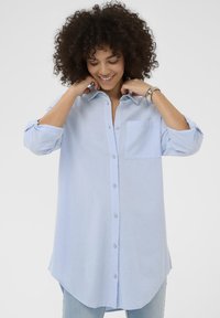 Vaaleansininen nappipaita, jossa on tasku, käännetyt hihansuut ja pyöristetty helma. Valmistettu pehmeästä kankaasta, rennolla istuvuudella.