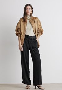 Satin bomber jakna v tan barvi z rebrastimi manšetami, bež bluzo in širokimi črnimi cargo hlačami. Model nosi črne sandale s trakovi in debelo ogrlico.