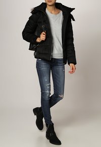 Sort polstret jakke med hætte, grå skjorte under, slidte blå jeans og sorte ankelstøvler med spænde detaljer.