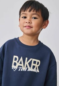 Marineblå sweatshirt med ribbet halsudskæring, der har "BAKER" skrevet med store cremefarvede bogstaver og "by Ted Baker" i mindre skrift nedenunder.