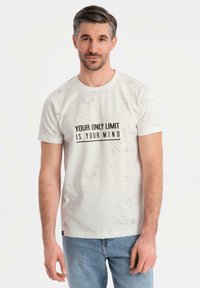 Camiseta de algodón blanca con un patrón moteado, que presenta un estampado en texto negro centrado: "TU ÚNICO LÍMITE ES TU MENTE." Cuello redondo, mangas cortas.