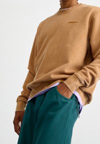 Felpa marrone con scollo tondo e logo ricamato. Abbinata a pantaloni turchese con coulisse. Tessuto liscio e morbido.