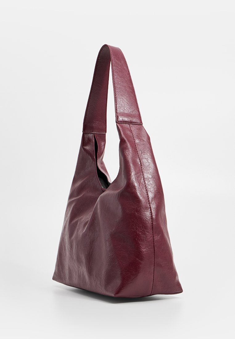 Bolso de hombro de cuero burdeos suave con una correa ancha y forma holgada, desestructurada, sobre un fondo blanco liso.