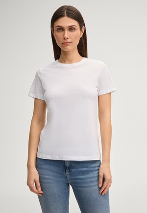 Femme aux cheveux bruns raides, portant un t-shirt blanc simple à manches courtes et un jean bleu, debout devant un fond neutre.