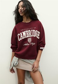 Kvinde med langt hår iført en burgunder University of Cambridge sweatshirt, stribet skjorte knyttet om livet, beige mini nederdel og gyldne ringe.