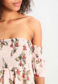 Top floreale scollato sulle spalle in rosa chiaro. Presenta un corpetto arricciato, maniche corte a palloncino e vivaci motivi floreali rossi e verdi.