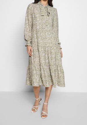 Kobieta w długiej, sięgającej kolan sukience w kwiaty z kołnierzykiem z kokardą i warstwową spódnicą, ubrana w białe sandały na obcasie z paskami.