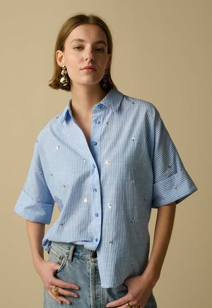 Woman wearing a blue striped short-sleeve shirt with small silver embellishments, denim jeans, gold earrings, and rings, standing with hands in pockets.