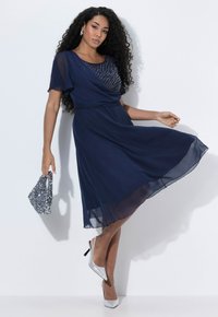 Navyfarbendes Chiffonkleid mit strukturiertem Oberteil und ausgestelltem Rock, kombiniert mit silbernen, glitzernden Absätzen und einer passenden Clutch.