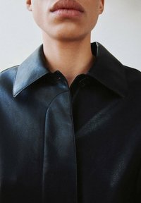 Czarna skórzana kurtka z dużym kołnierzem, gładką fakturą i zapięciem na guziki. Widok z bliska podkreśla szczegóły szwów.