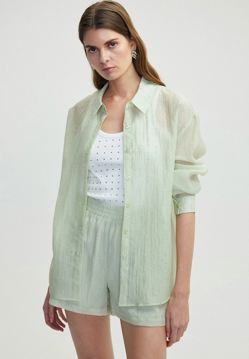 Camisa botón ligera de color verde claro, semitranslúcida, con un ajuste relajado y una textura sutil, llevada sobre un top blanco con puntos negros y pantalones cortos a juego.
