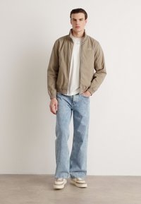 Jachetă bomber de culoare maro deschis, cu guler în picioare, asociată cu blugi denim lejeri de culoare albastră deschisă și adidași bej. Tricou alb simplu dedesubt.