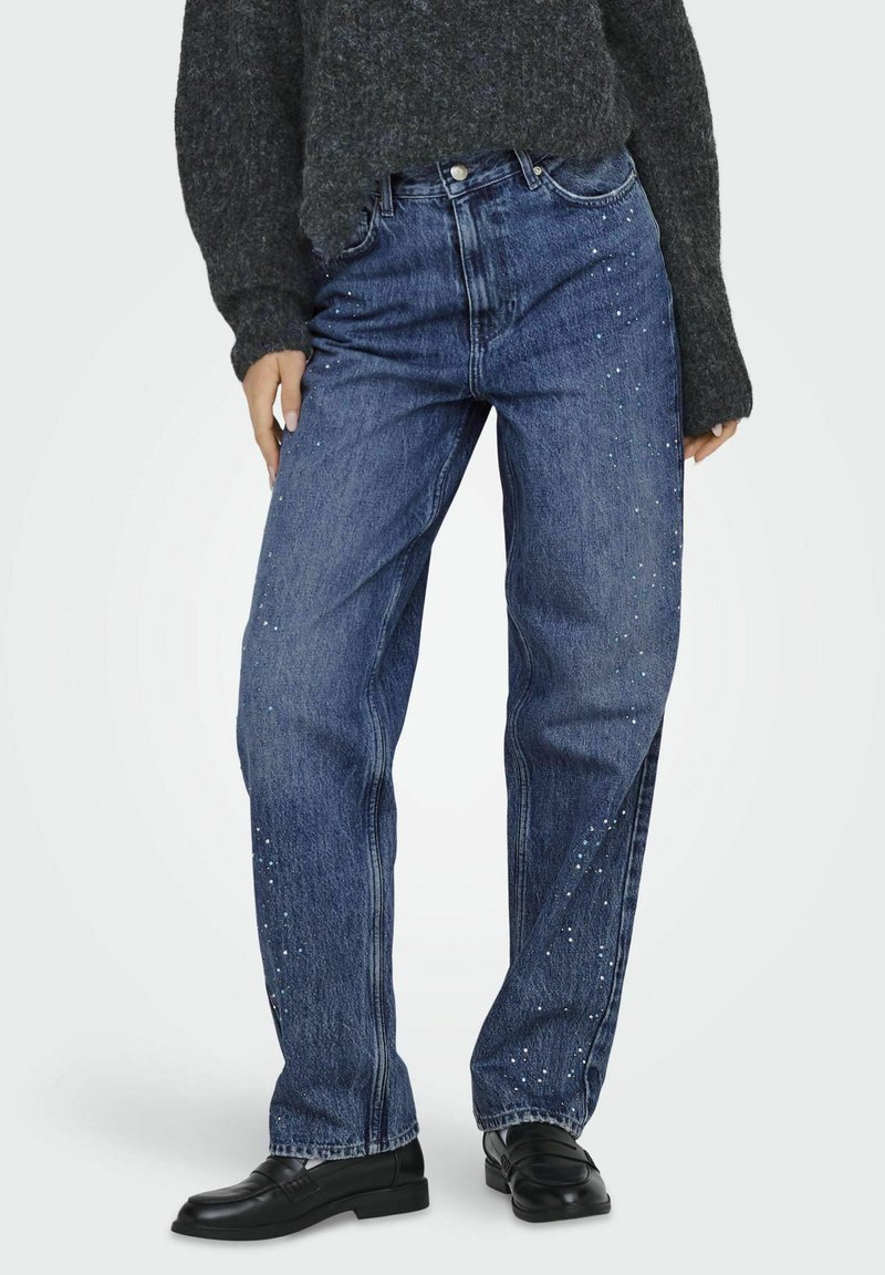 Jeans din denim albastru cu o croială relaxată, prezentând embellisamente împrăștiate pe cusăturile exterioare, purtate împreună cu un pulover texturat închise la culoare.