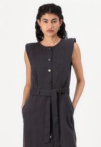 Vestido cinza escuro sem mangas com frente abotoadas, cintura cingida por um cinto, bolsos laterais e uma textura de tecido suave. Design simples e estruturado.