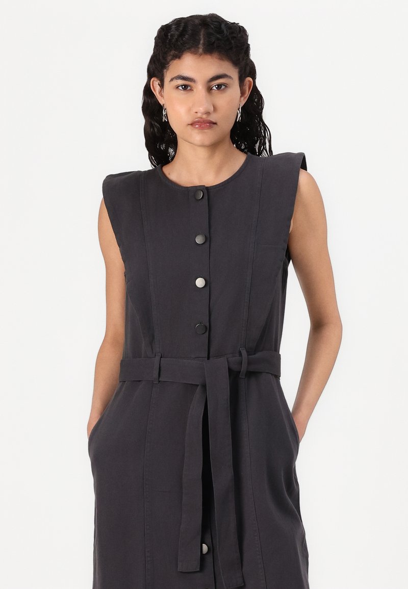Ärmelloses dunkelgraues Kleid mit Knopfleiste auf der Vorderseite, tailliertem Gürtel, Seitentaschen und glatter Stoffstruktur. Einfaches, strukturiertes Design.