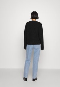 Kobieta z krótkimi czarnymi włosami stoi zwrócona twarzą do ściany, ubrana w czarny sweter z długim rękawem, jasnoniebieskie dżinsy i czarne buty na obcasie.