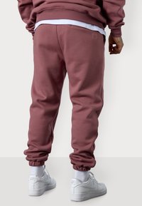 Burgunderfarbene Sweatpants mit elastischen Bündchen, seitlichen Taschen und dezentem Branding. Getragen mit weißen Sneakers und Knöchelsocken. Glatte Stofftextur.