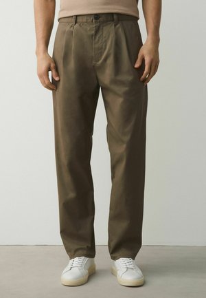 Vyras dėvi alyvuogių žalios spalvos tiesaus kirpimo kelnes ir baltus sportbačius, stovintis priešais paprastą šviesų sienos foną.